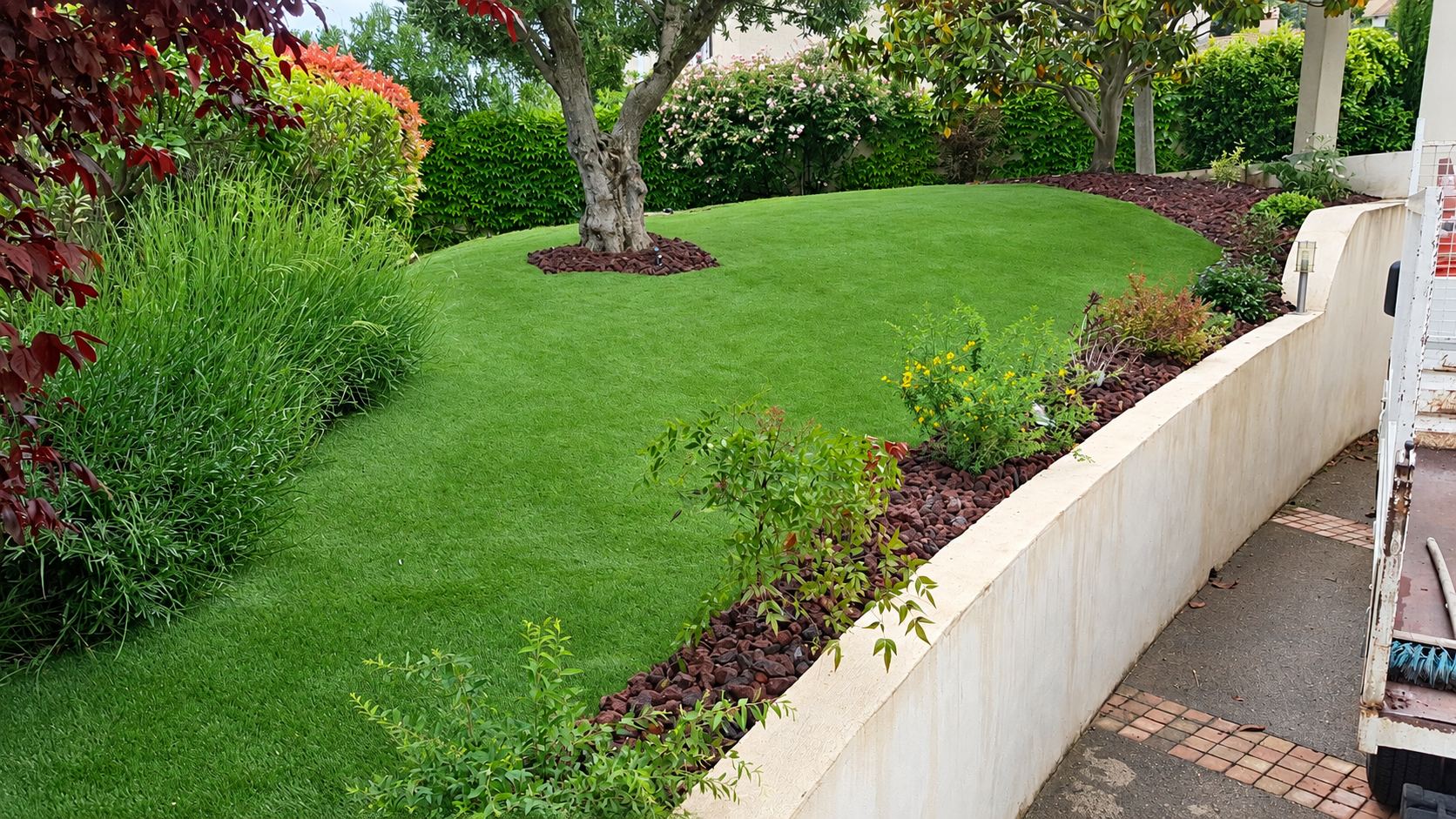 Jardin en pente avec pelouse synthétique, olivier et massifs paillés