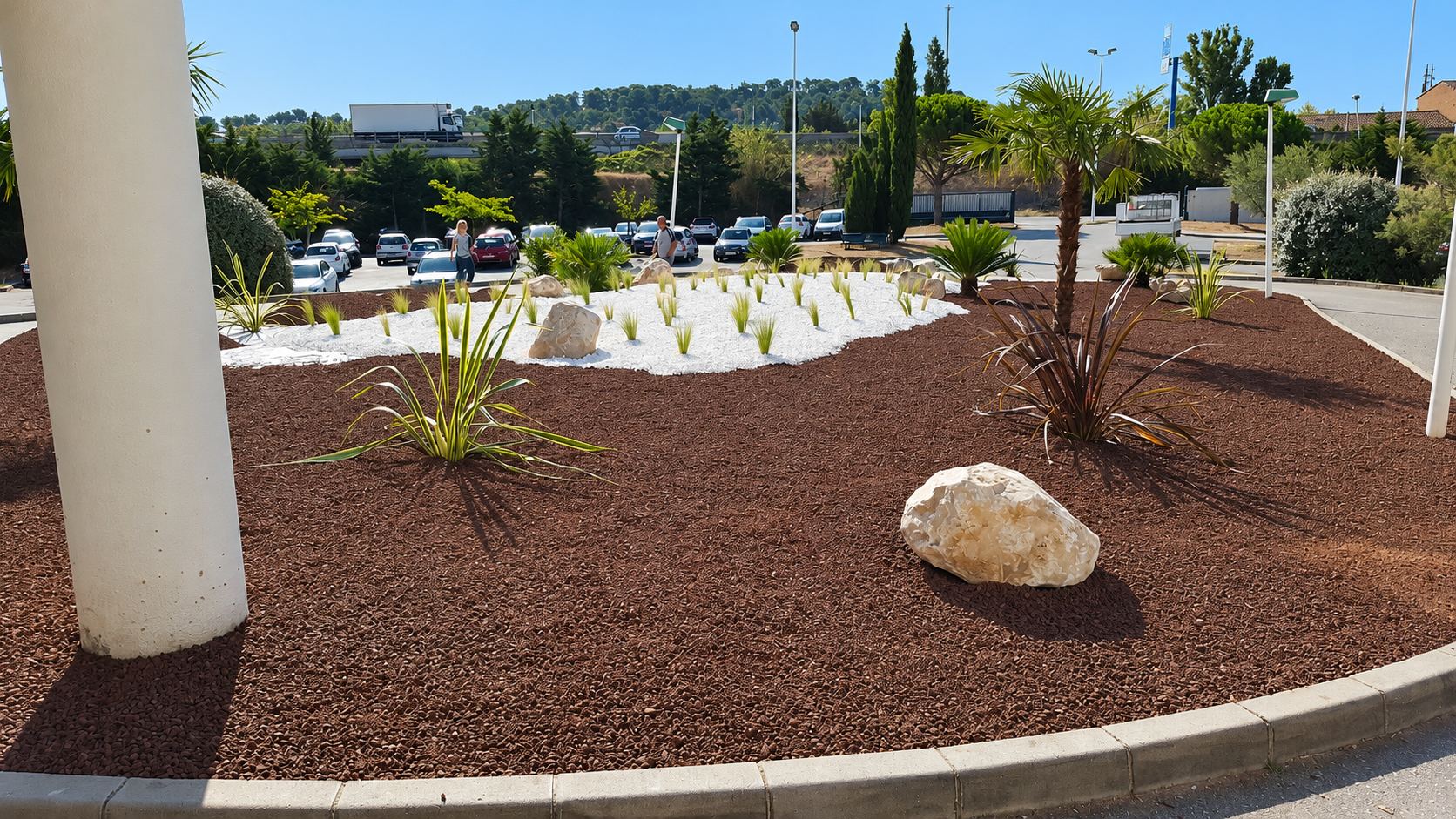 Massif paysager avec paillage rouge, galets blancs, palmier et rochers près d'un parking
