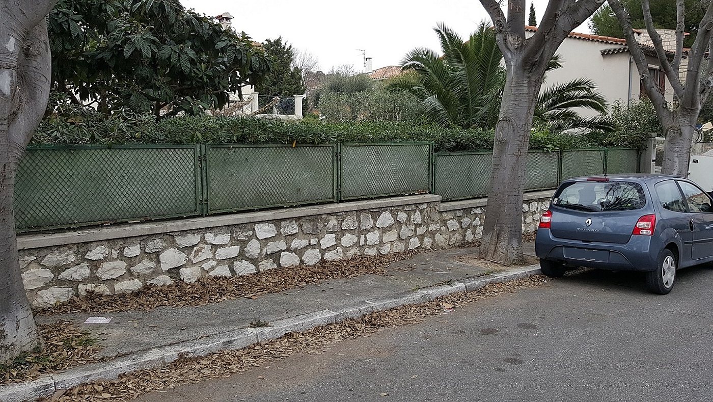 Muret en pierres surmonté d'un grillage vert le long d'une rue